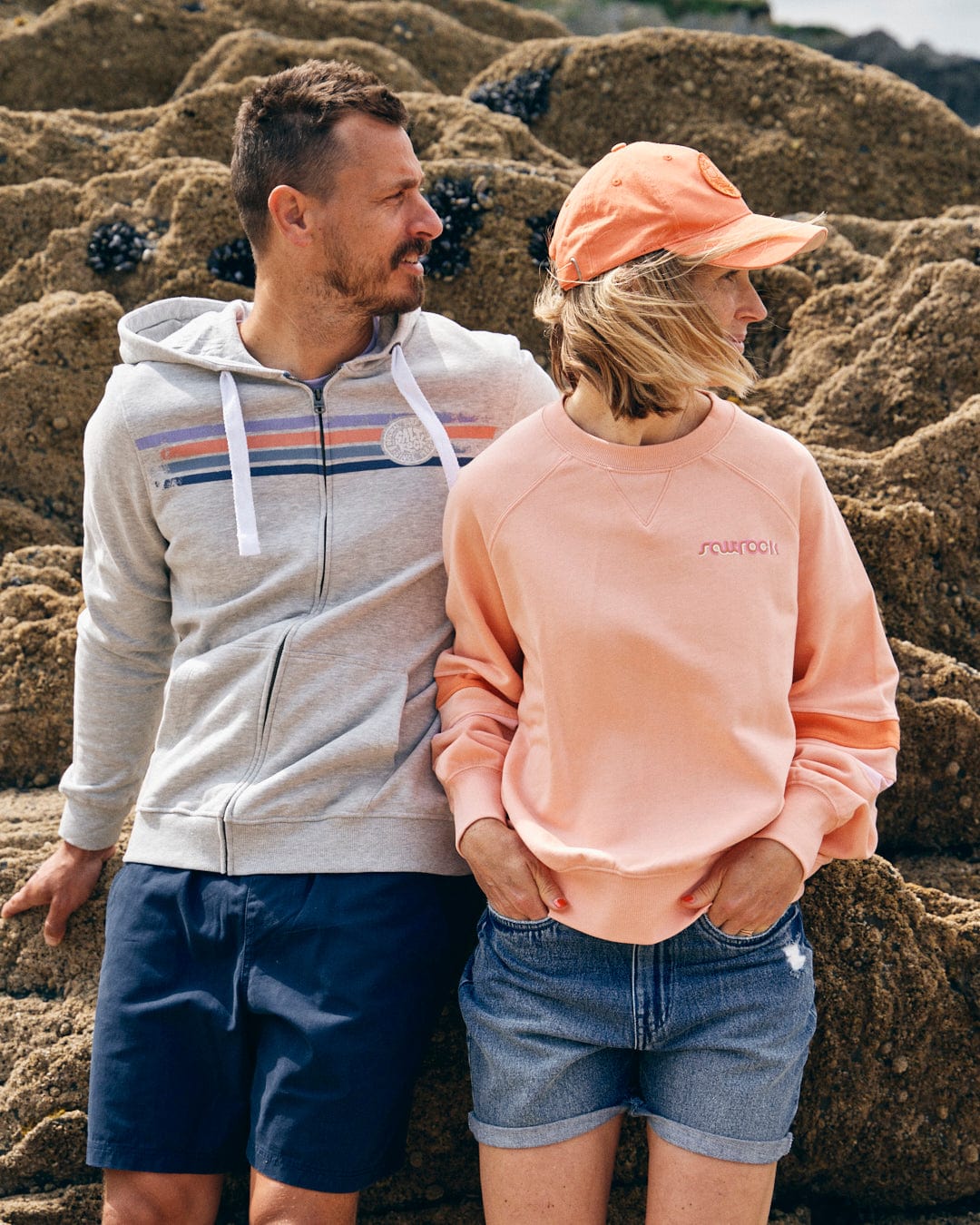 A man and woman are standing against a rocky background. The man is wearing a grey hoodie with Saltrock branding and blue shorts, while the woman is sporting the "Retro Ribbon Block" oversized peach sweatshirt from Saltrock, denim shorts, and an orange cap.