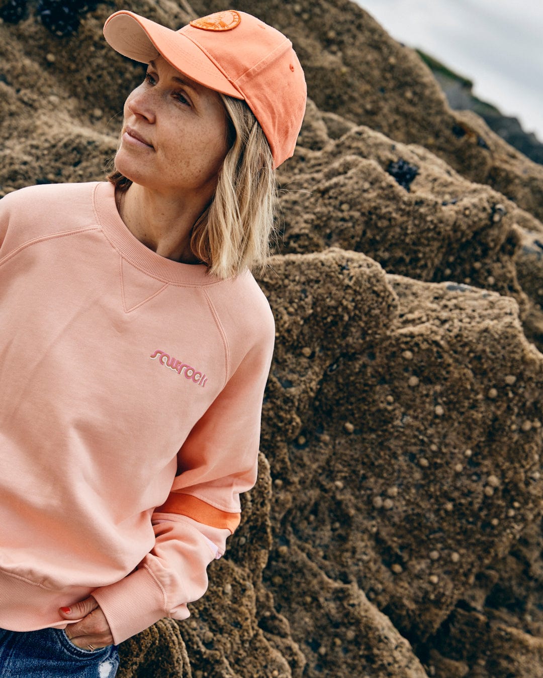 Woman wearing the Retro Ribbon Block - Womens Sweatshirt in peach from Saltrock, paired with a matching Saltrock-branded baseball cap, standing against a rocky background.