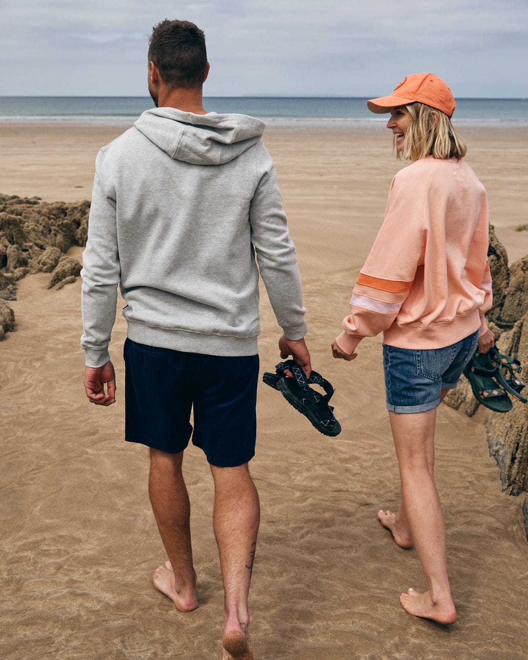 Two people walking barefoot on a beach, holding their sandals. The person on the left is in a grey cotton hoodie and shorts, while the person on the right wears an oversized Saltrock-branded Retro Ribbon Block Women's Sweatshirt in Peach, denim shorts, and an orange cap.