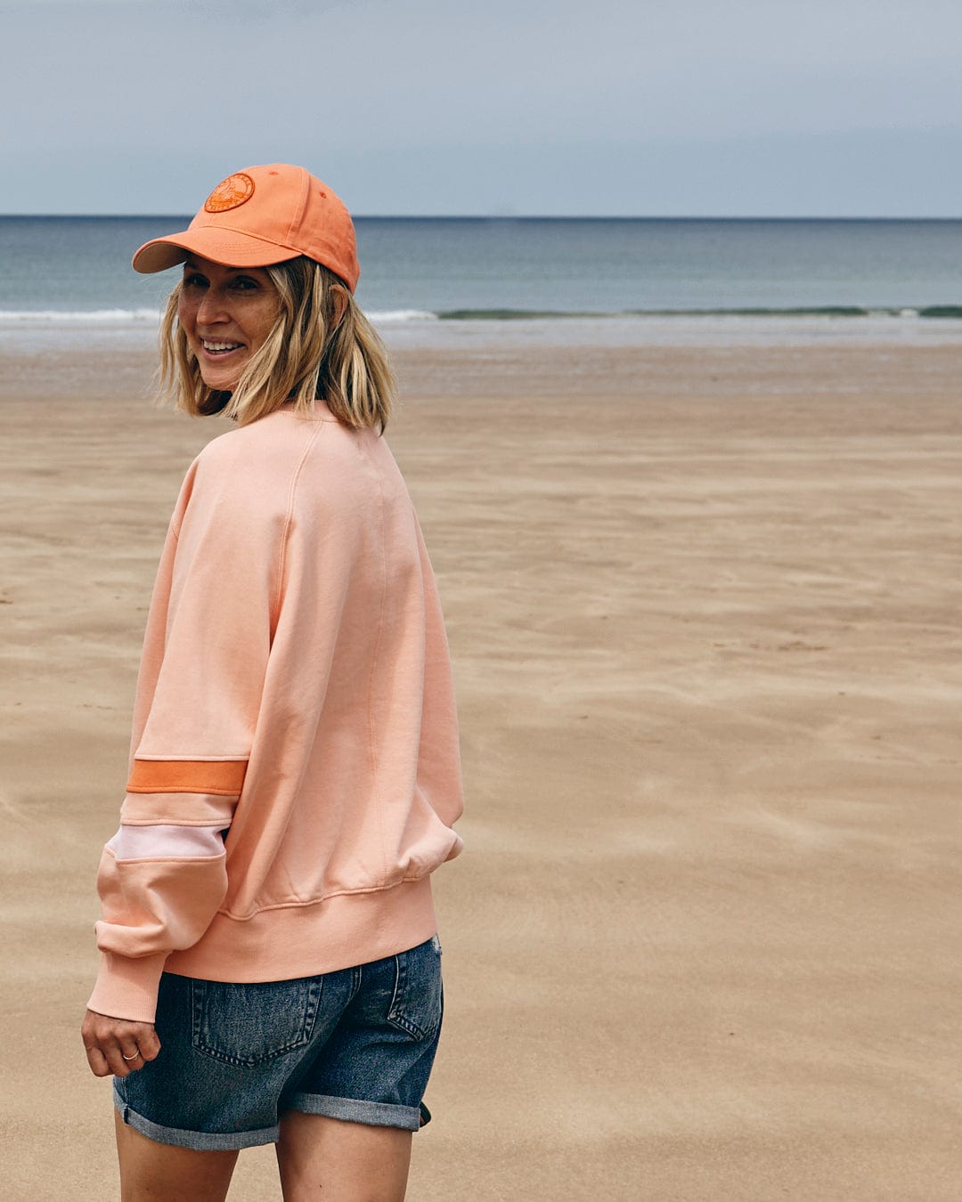 A person in a peach-colored Retro Ribbon Block women's sweatshirt by Saltrock, paired with denim shorts, stands on a beach, facing away while smiling over their shoulder. The Saltrock branding is visible as the sea and sky blend into the background.