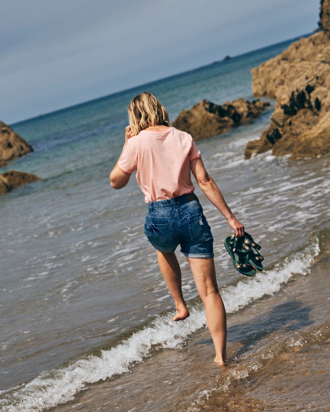 A person in a Retro Ribbon Womens Short Sleeve T-Shirt in Peach by Saltrock and denim shorts walks barefoot at the edge of the sea, holding sandals, with rocks and the horizon in the background.