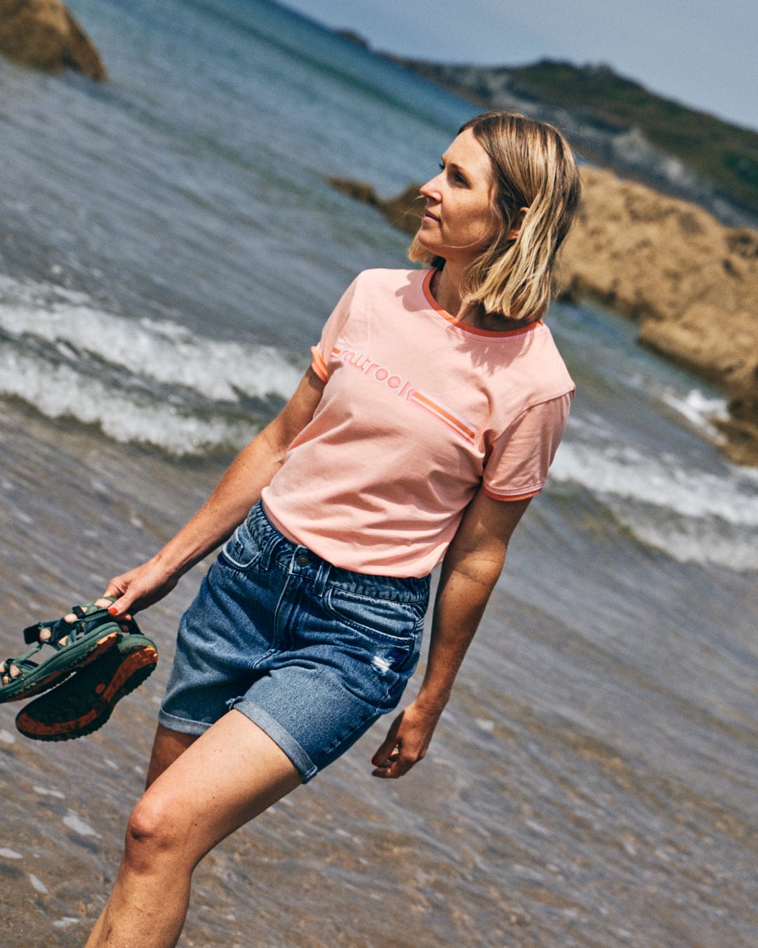 A woman in a Saltrock Retro Ribbon - Womens Shorts Sleeve T-Shirt in peach and denim shorts walks barefoot on a sandy beach, holding Saltrock-branded sandals in her hand. Rocky formations and waves are visible in the background.