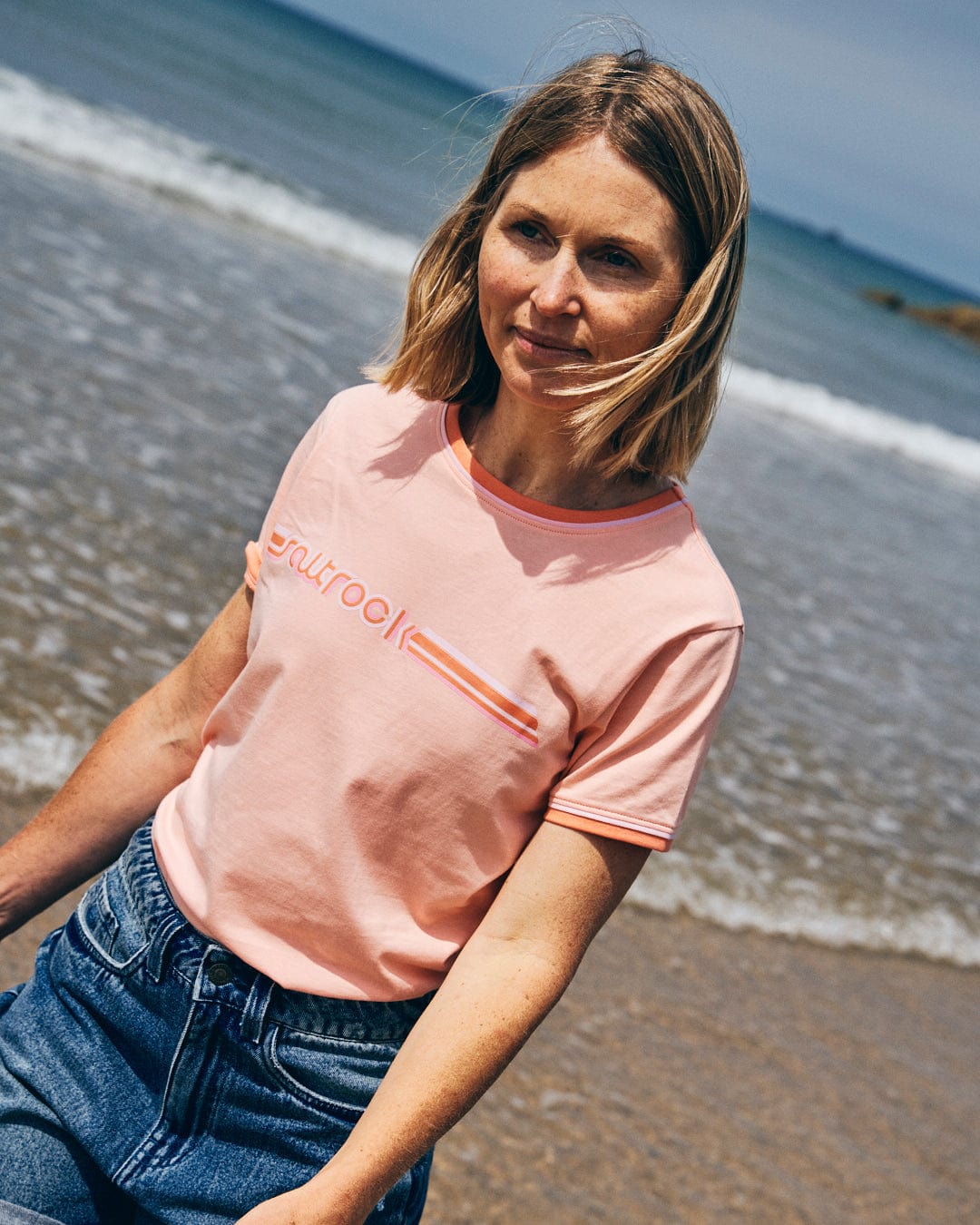 A person with shoulder-length hair wearing a Saltrock Retro Ribbon Women's Short Sleeve T-Shirt in Peach and blue jeans walks along a sandy beach with waves in the background.
