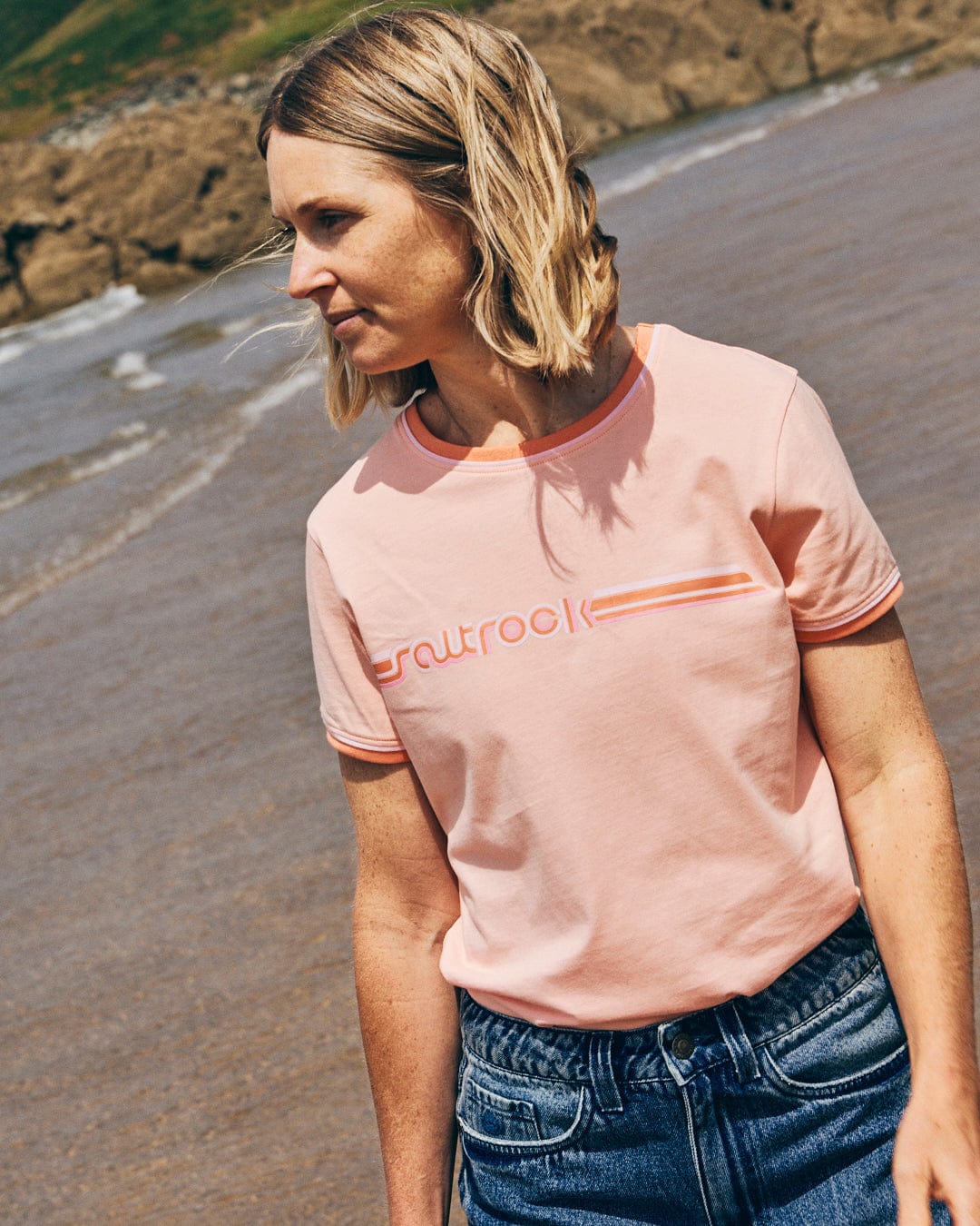 A person with short blonde hair is wearing the Retro Ribbon women's short sleeve T-shirt in peach and jeans, standing on a sandy beach with water and rock formations in the background, showcasing Saltrock branding.