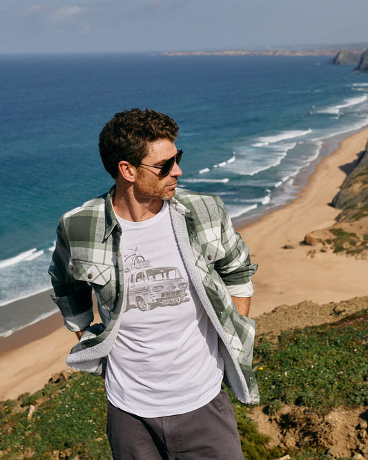A man, wearing a Saltrock Re-Wild - Mens Short Sleeve T-Shirt in White, stands on a hill overlooking the ocean.