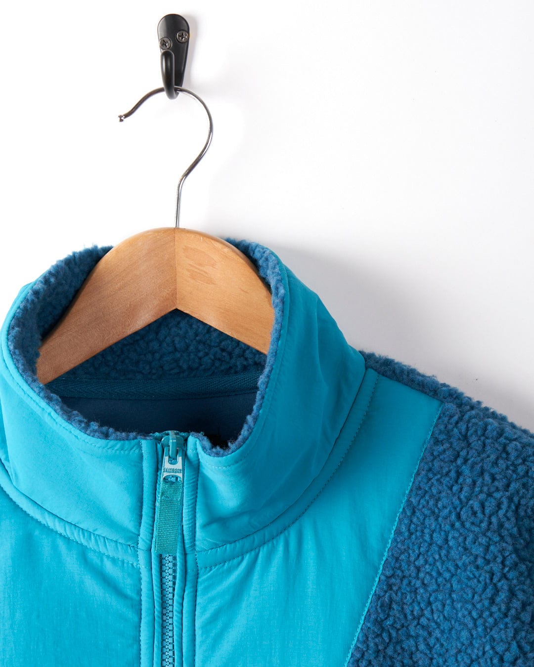 The Saltrock Robyn Women's Fleece in blue, featuring cosy fleece material and Saltrock branding, hangs on a wooden hanger mounted on a wall hook.