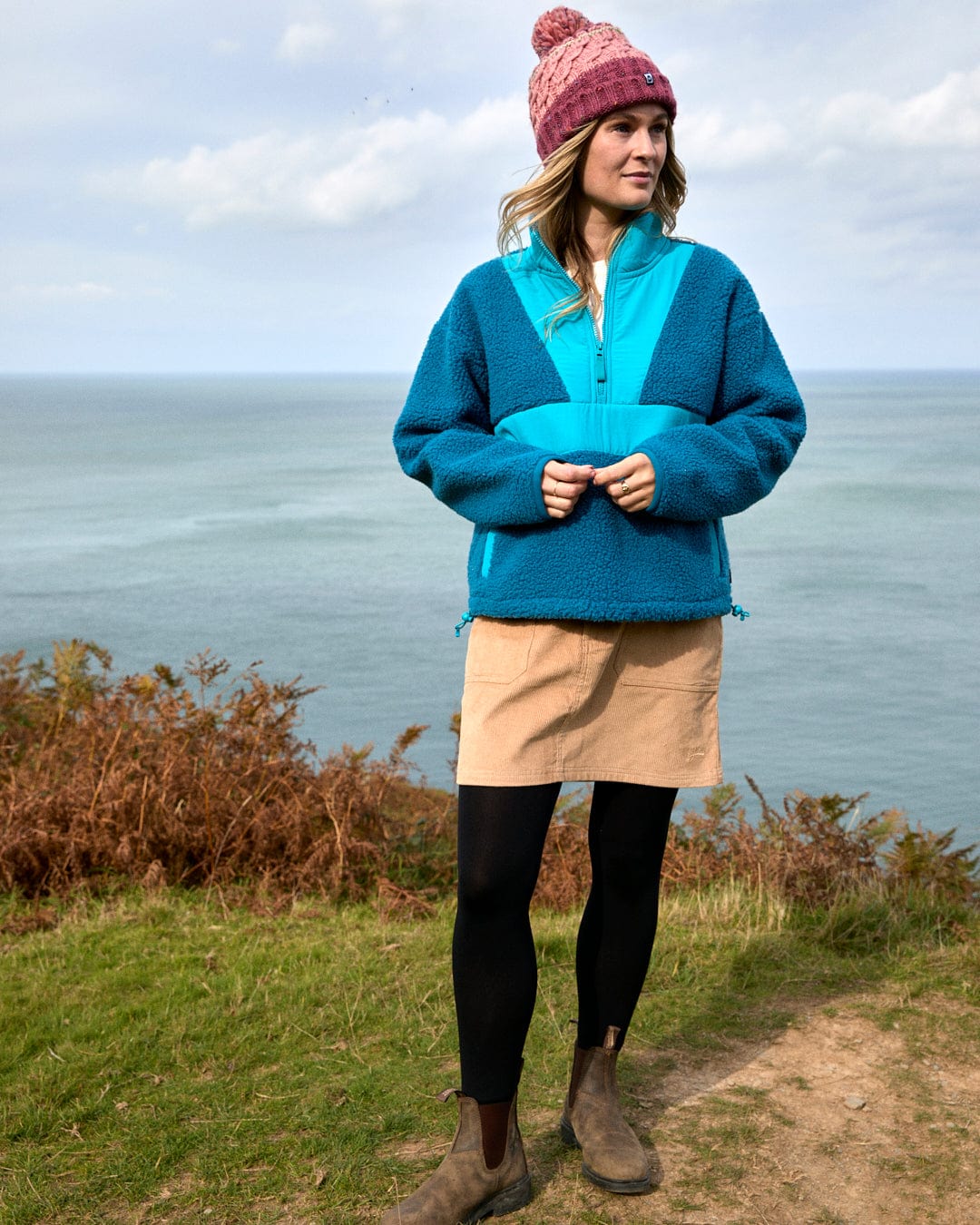 A person wearing a pink knit hat, tan skirt, black tights, brown boots, and the Saltrock Robyn Women's Fleece in blue stands on a grassy path by the sea.