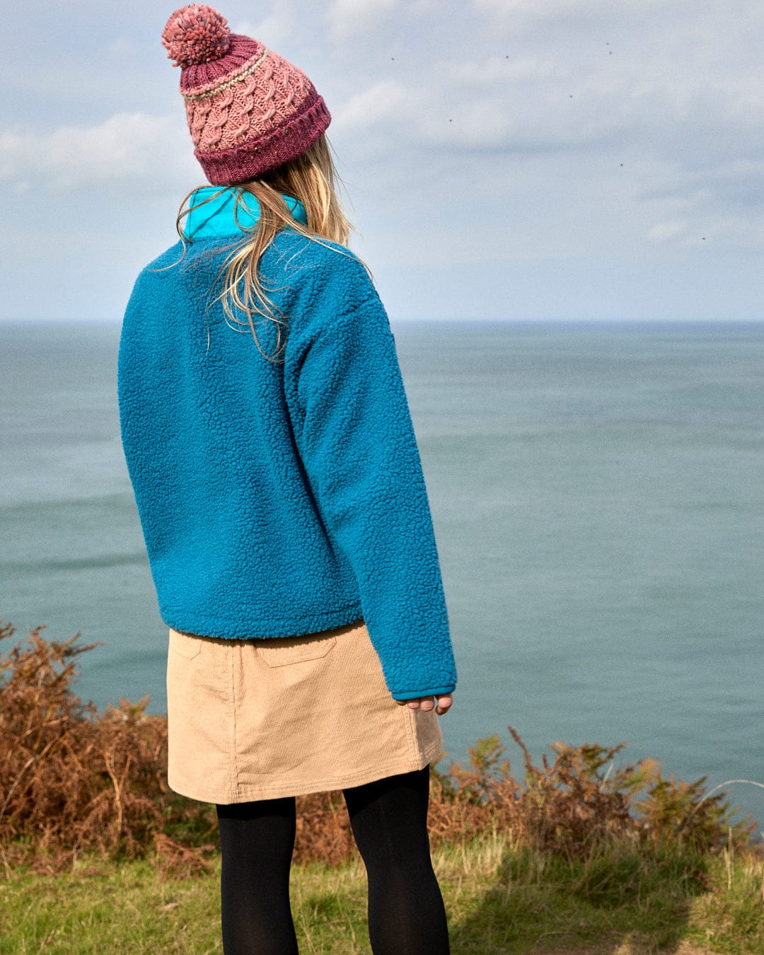 A woman wearing the Saltrock Robyn Women's Fleece in blue stands on a grassy cliff overlooking the sea, paired with a beige skirt, black tights, and pink knit hat under a cloudy sky.