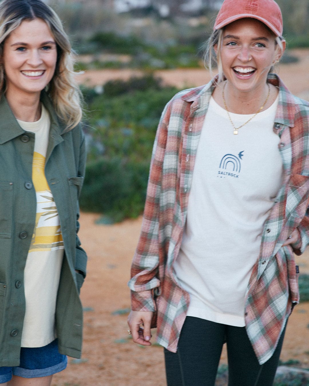 Two smiling women standing outdoors, one wearing a Saltrock Rosalin Womens Long Sleeve Shirt in orange/green with contrasting buttons over a graphic tee and the other in a green jacket and striped Longline shirt.