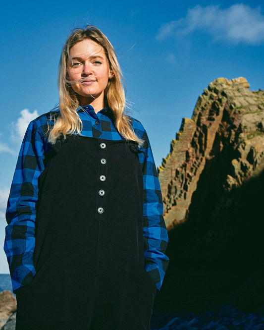 A woman in Saltrock Rosalin - Womens Check Shirt - Blue/Black overalls standing in front of rocks.