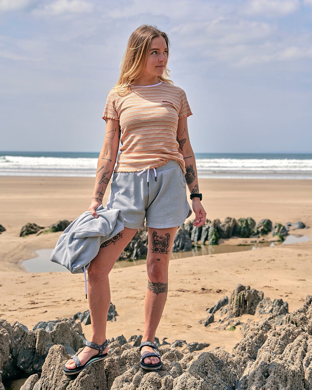 A woman wearing Rosalin - Womens Sweat Shorts - Light Grey from Saltrock, and a tee shirt standing on rocks.