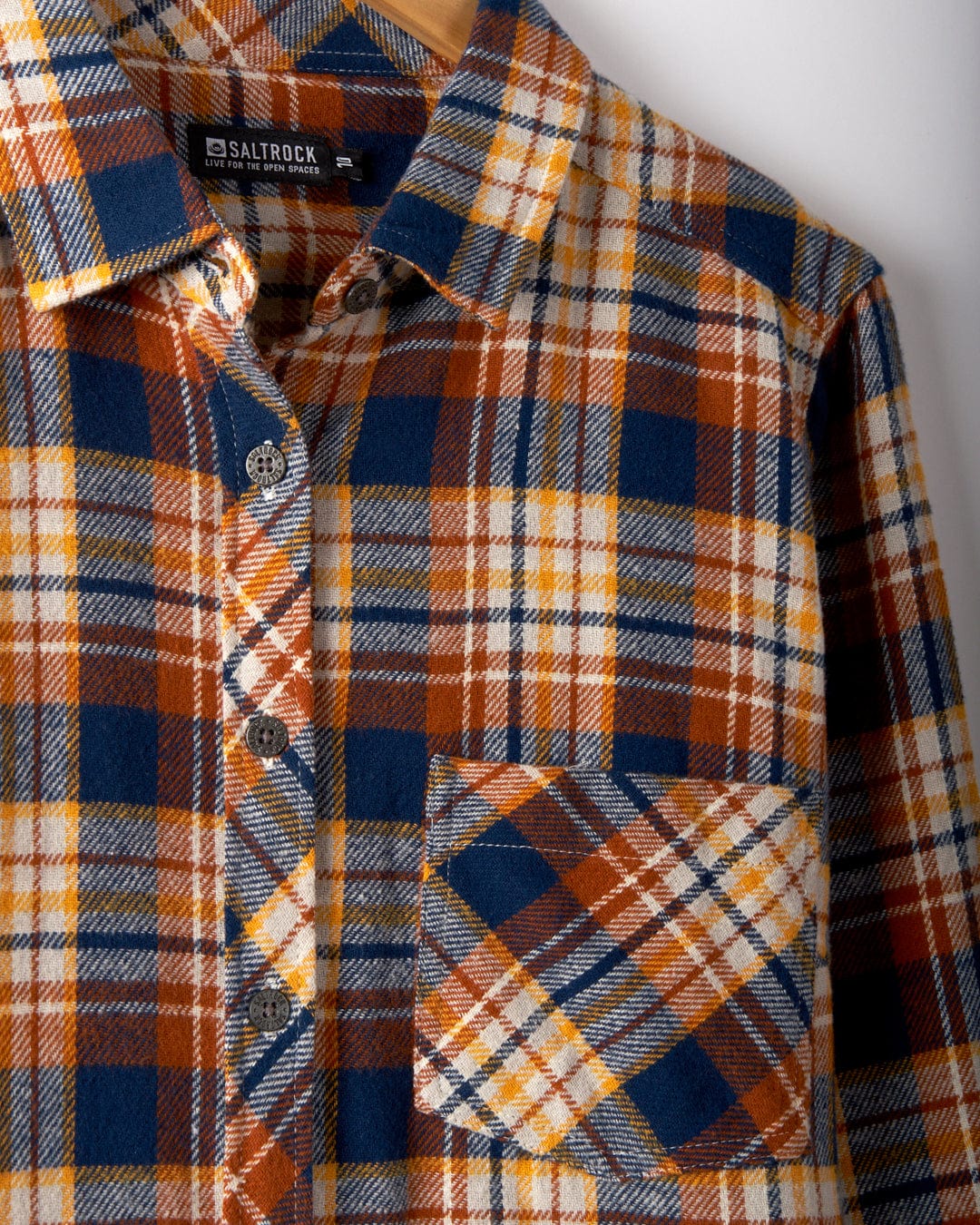 A close-up of Saltrock's Rosalin Women's Check Shirt in Burnt Orange hangs on a hanger. Made from soft brushed cotton, it features a blue, white, orange, and yellow pattern with a chest pocket and stylish metal buttons.