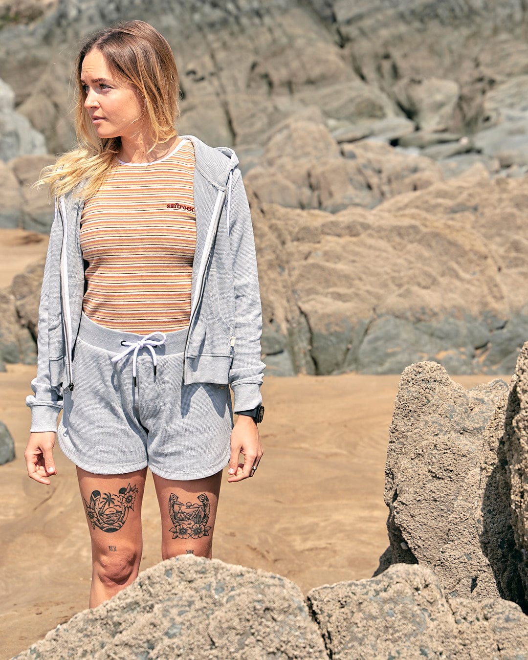 A woman with tattoos standing on a rocky beach wearing the Saltrock Rosalin - Womens Zip Hoodie - Light Grey.