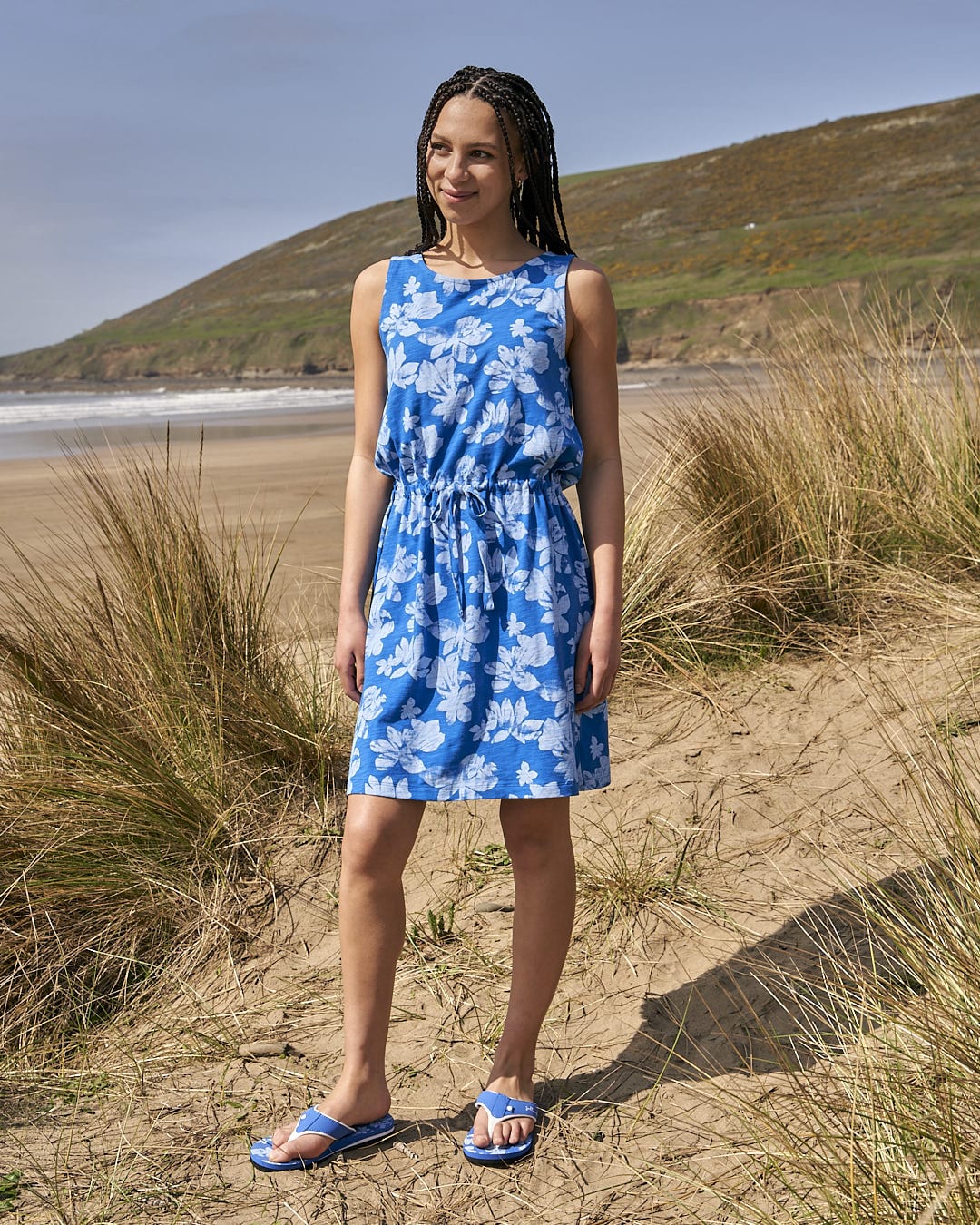 A woman in a Saltrock Floral - Womens Tie Vest Dress - Blue standing on the beach.