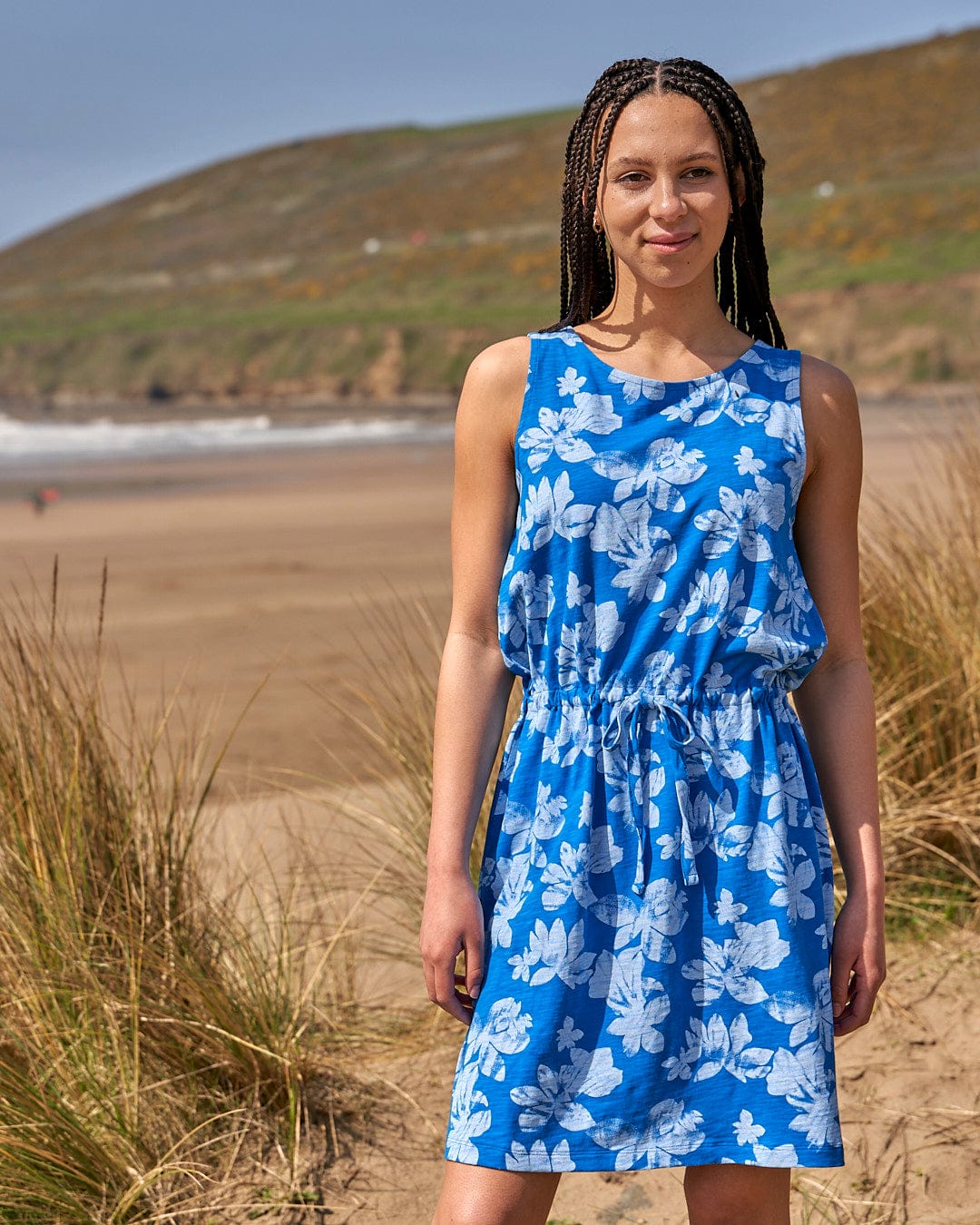 A woman in a Saltrock Floral - Womens Tie Vest Dress - Blue standing on a beach.