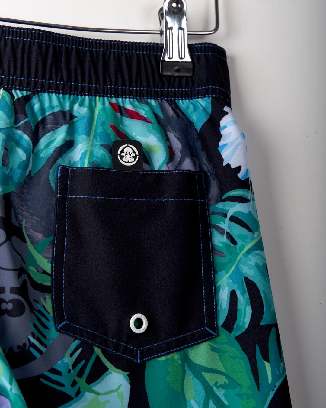 Close-up of a pair of Rumble - Kids Recycled Boardshorts - Multi with a tropical leaf design, crafted from recycled materials. Featuring a black pocket with blue stitching and a small skull logo, these 4-way stretch trunks from Saltrock are shown hanging on a clip.