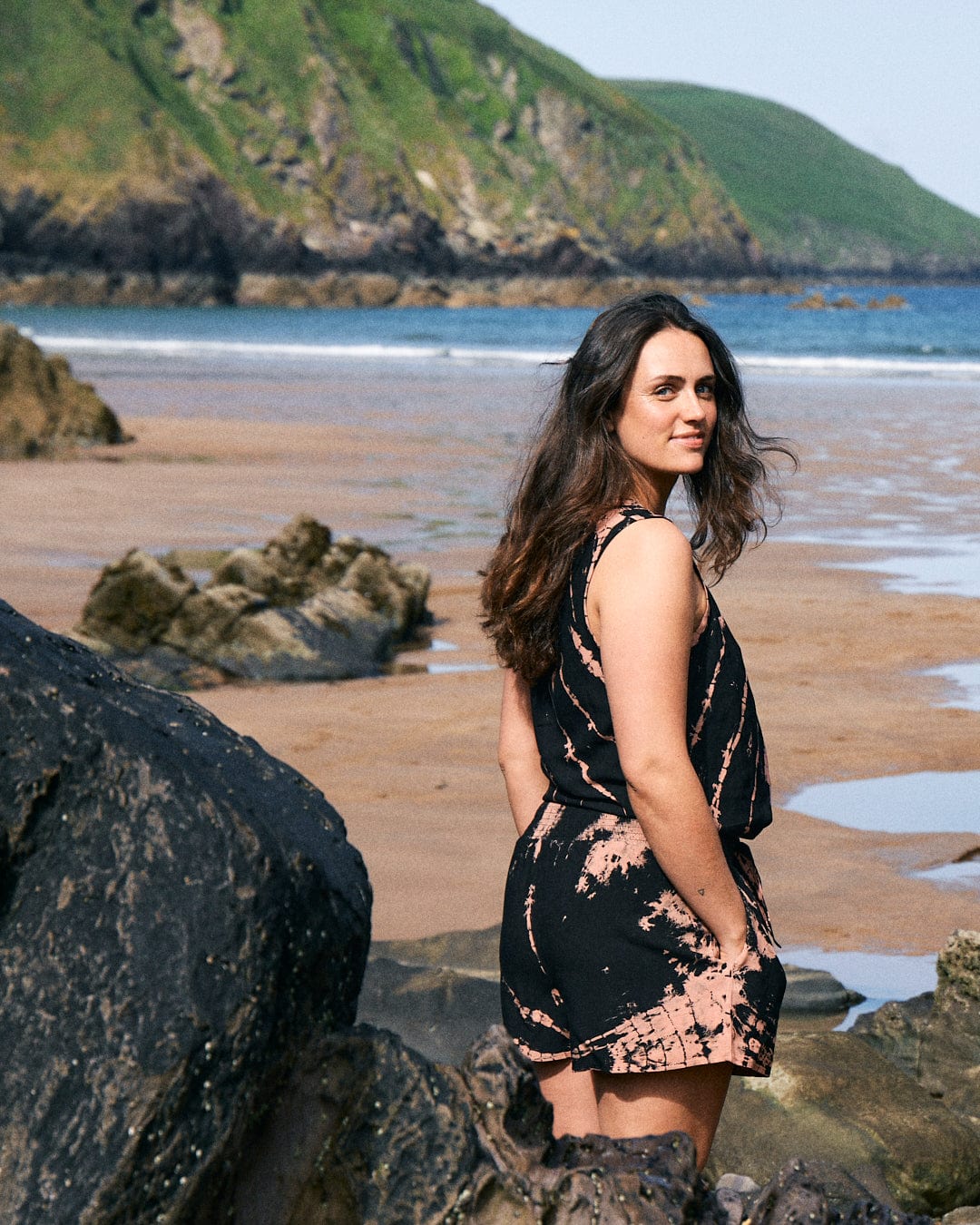 A woman wearing a Saltrock Saffron - Womens Tie Front Vest - Black stands on a rocky beach. The shoreline, waves, and green cliffs are visible in the background. She has long, dark hair and is looking back over her shoulder.