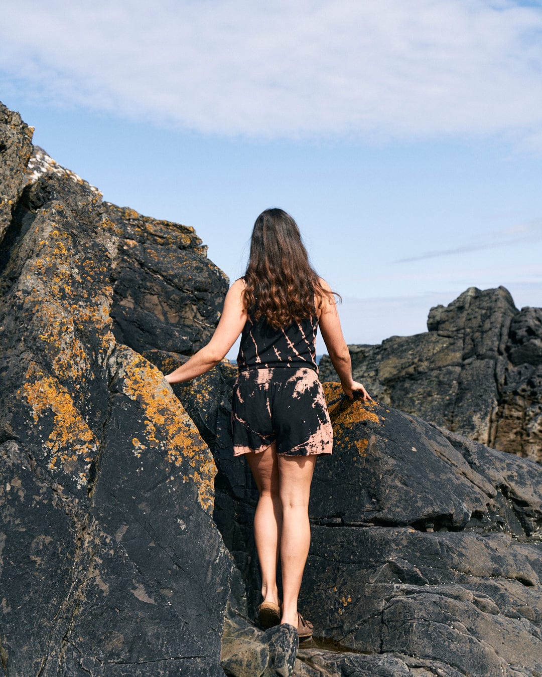 A woman with long hair stands on a rocky terrain under a partly cloudy sky. She is wearing the Saltrock Saffron - Womens Tie Front Vest - Black, facing away from the camera, with one hand touching the rocks. The lightweight material of her clothes allows for ease of movement as she explores the rugged landscape.