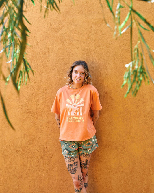 A person in a Saltrock Sunshine - Recycled Womens Cropped T-Shirt - Orange with Saltrock branding and patterned shorts stands against an orange wall. They have tattoos on their legs and arms, while greenery hangs from the top of the image, casting a mystic dessert sunset vibe.