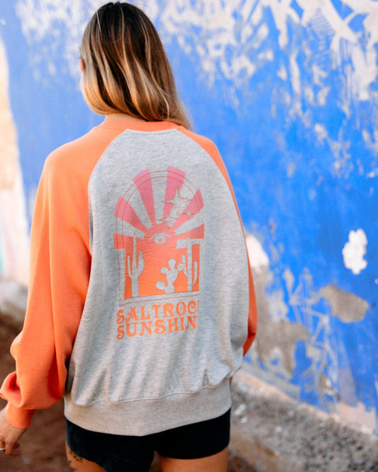 Woman wearing a gray and orange Saltrock hoodie with "Saltrock Sunshine" graphic and raglan sleeves, standing in front of a blue weathered wall.