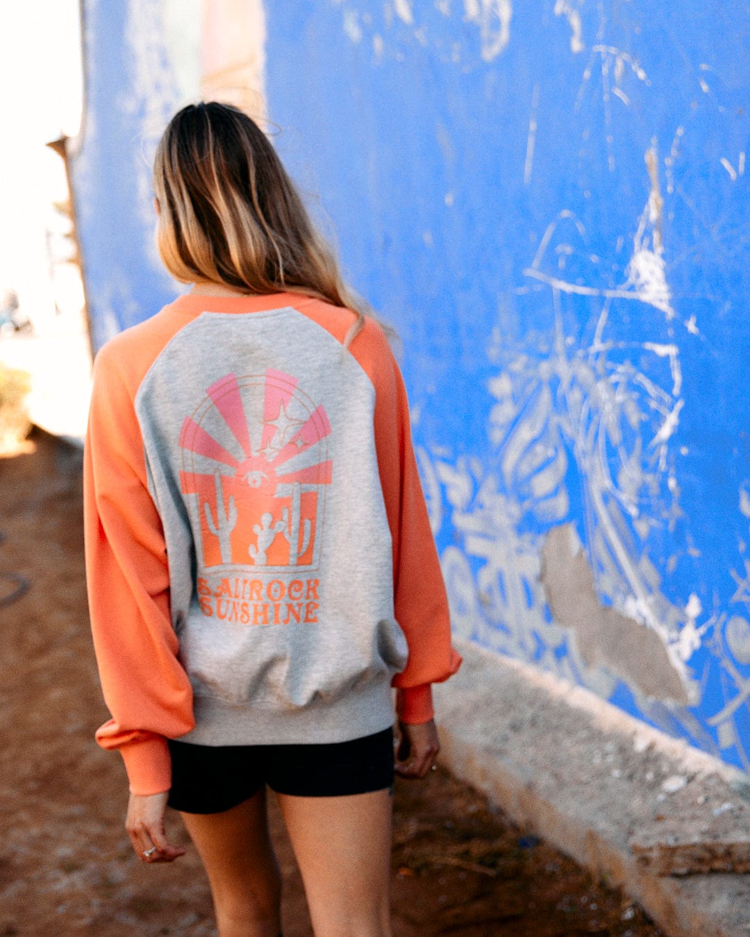 Woman in a Saltrock Sunshine - Womens Raglan Sweat - Grey Marl hoodie with raglan sleeves walking past a blue wall with scraped paint.