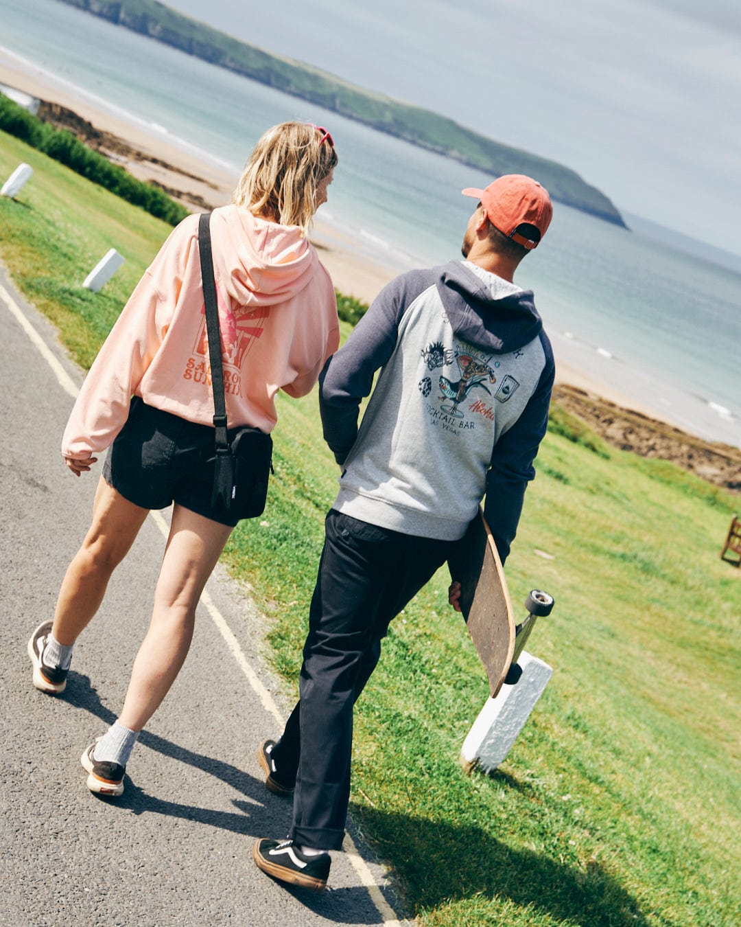 Two people walk on a paved path near a beach. One person holds a skateboard, and both are dressed casually in relaxed fit hoodies and shorts, facing away from the camera. One sports the Saltrock Sunshine Women's Pop Hood in Peach with mystical ombre designs from Saltrock. The sea and distant land are visible.