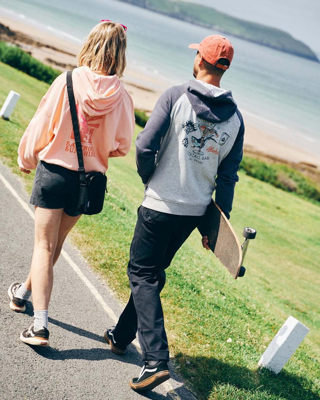 Two people walk on a beachside path, one carrying a skateboard. Both wear casual clothing with relaxed fit hoodies and sneakers. The sea and green landscape are visible in the background, with one sporting an embroidered Saltrock hoodie featuring a mystical ombre design, while the other is wearing the Saltrock Sunshine - Women's Pop Hood in Peach.