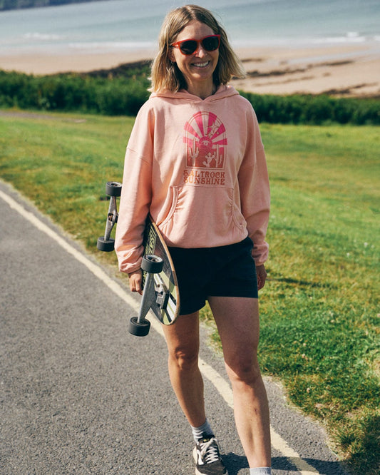 A person walking on a path near the beach is holding a skateboard and wearing Saltrock Sunshine's women's relaxed-fit pop hood in peach, along with sunglasses.