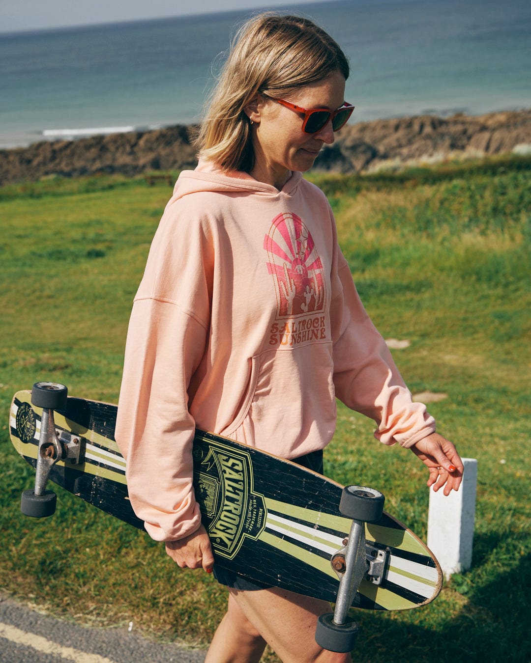 A person wearing the Saltrock Sunshine - Womens Pop Hood in Peach and sunglasses walks along a road carrying a longboard skateboard with a beach and ocean in the background.