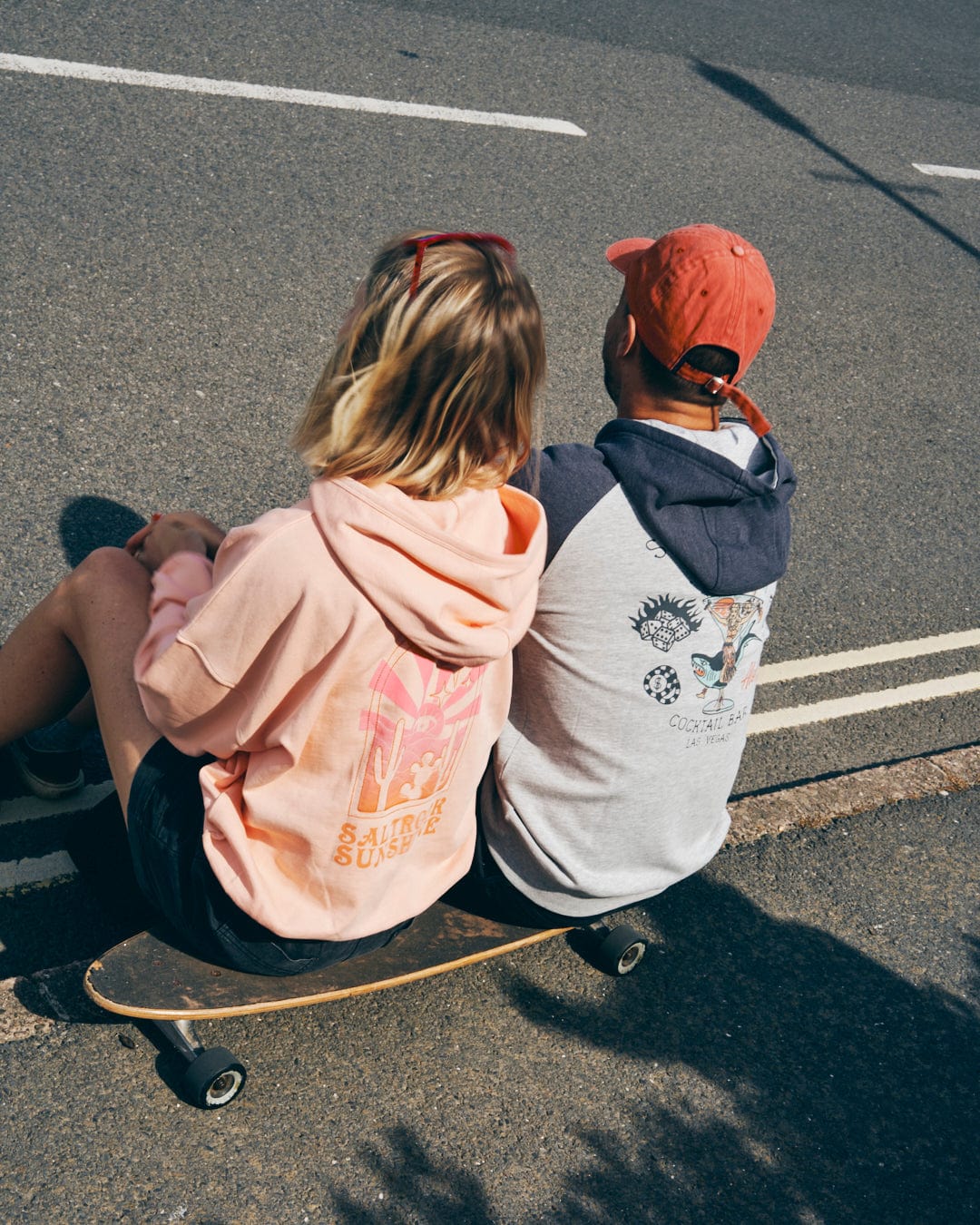 Two people sit on a skateboard on a road, embracing the relaxed fit of their casual hoodies and caps, one clad in a Saltrock Sunshine women's pop hood in peach and the other in a blue Saltrock mystical ombre hoodie, both facing away from the camera.