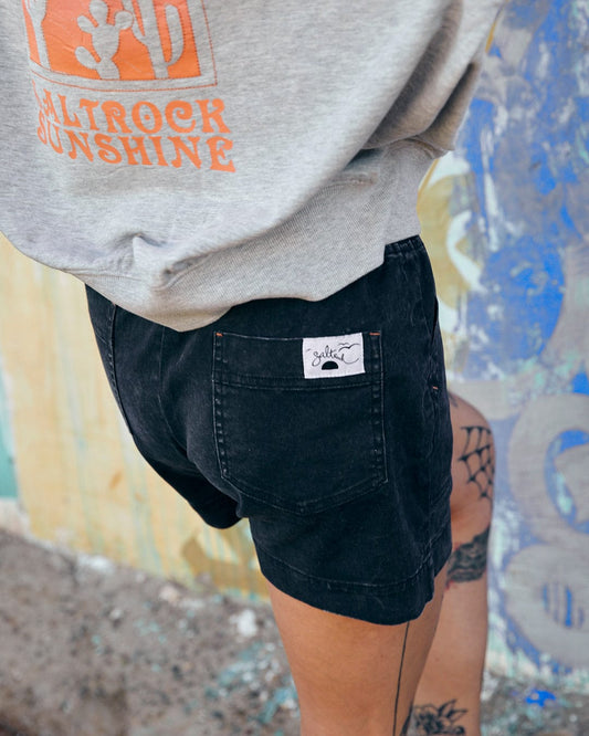 Person wearing a gray sweatshirt with an orange graphic and Saltrock Santano - Womens Short - Washed Black with a white tag on the pocket, standing with a tattoo visible on their leg.