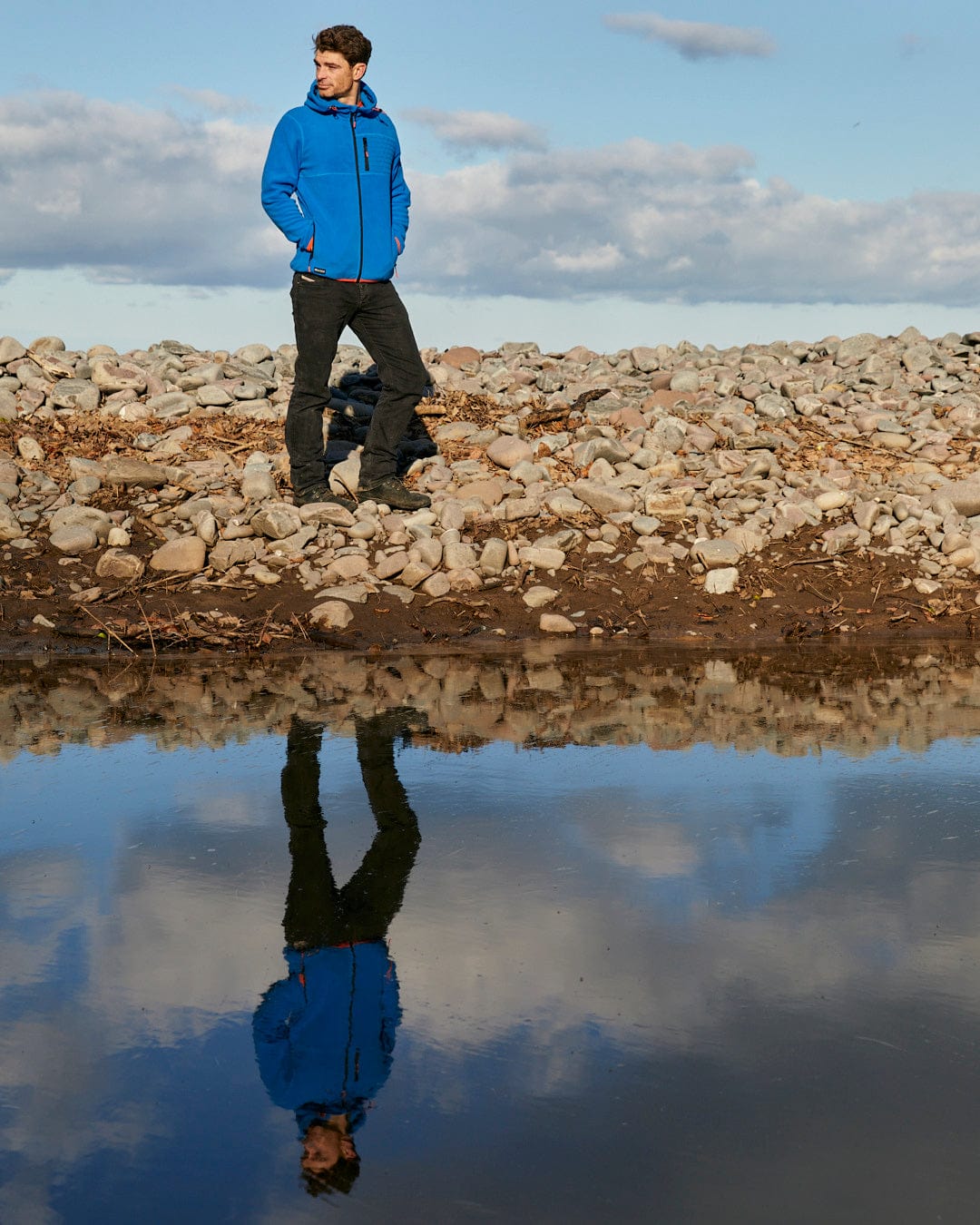 A man in a blue jacket showcasing Saltrock's Senja - Mens Fleece Hoodie - Blue while standing next to a pond.