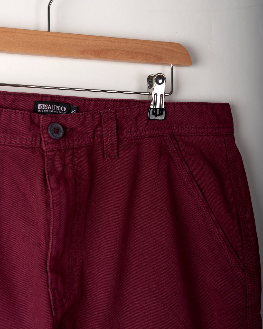 A pair of burgundy chino shorts with a relaxed fit hangs on a wooden hanger against a white background. The 100% cotton shorts have a Saltrock label indicating size 34.