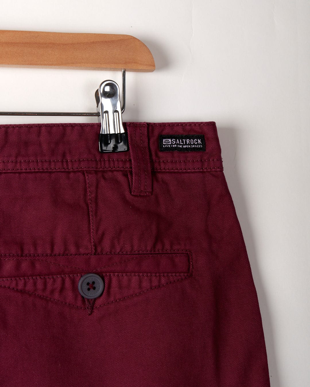 Close-up of the Sennen men's burgino chino shorts hanging on a wooden hanger with a metallic clip. These 100% cotton shorts feature a visible back pocket with a button and a small label reading "Saltrock" above it.