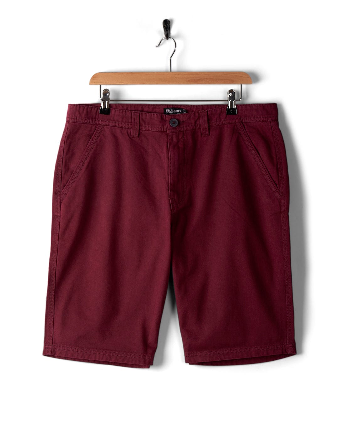 A pair of Sennen men's chino shorts in burgundy by Saltrock, made of 100% cotton fabric, displayed on a wooden hanger against a white background.