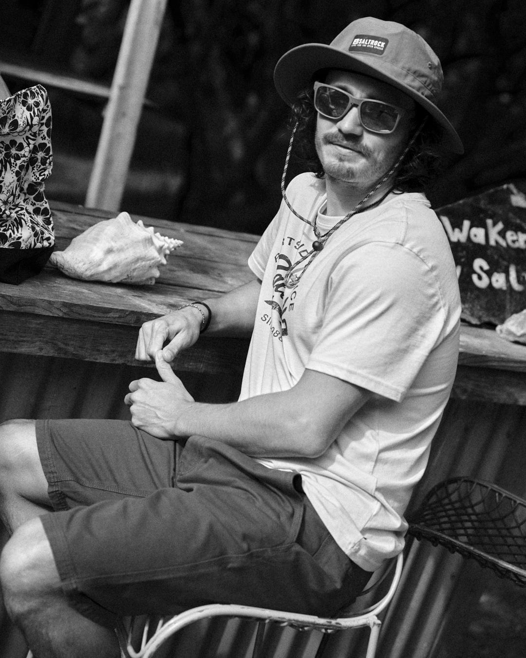 A person wearing a hat and sunglasses sits on a chair by a table with a shell and a bag, dressed in Saltrock's Sennen - Mens Chino Short in Burgundy. There is a sign behind them. Black and white image.
