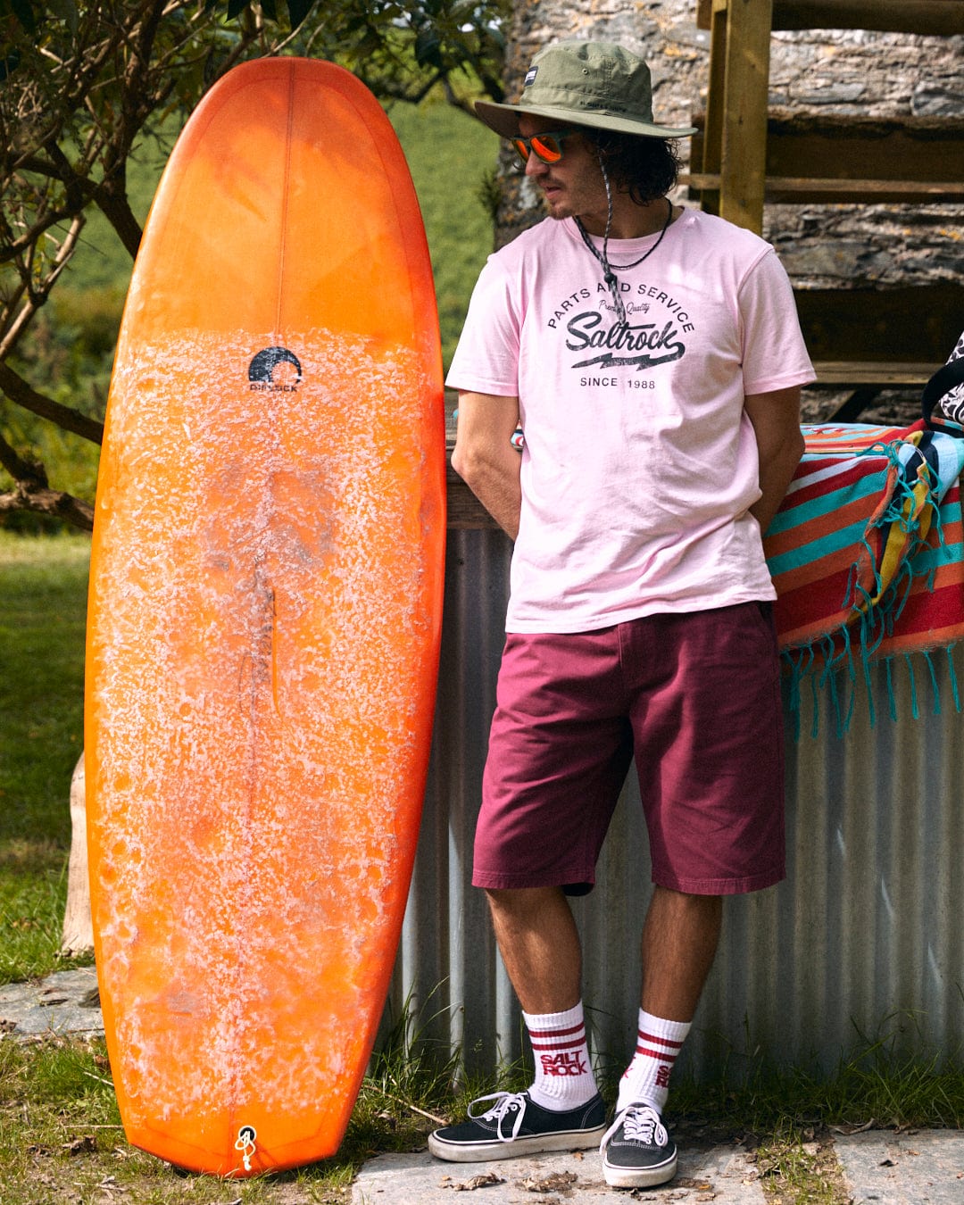 A person in a pink t-shirt and Saltrock's Sennen Men's Chino Short in burgundy, both made of 100% cotton, stands beside an orange surfboard, wearing sunglasses and a green hat.