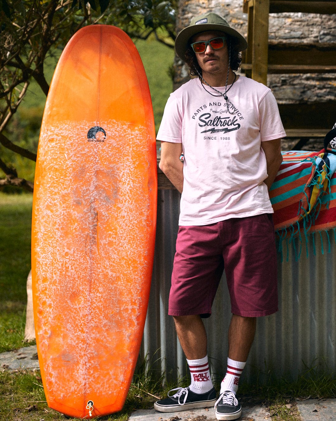 A person wearing a 100% cotton t-shirt, a pair of Saltrock Sennen Men's Chino Shorts in Burgundy, a hat, and sunglasses stands next to an orange surfboard that is leaning against a metal structure outdoors.