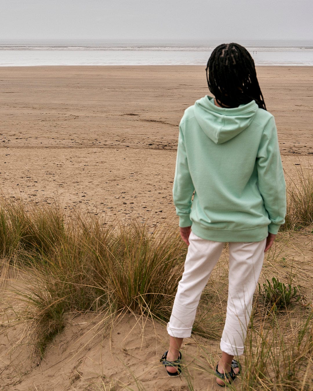 A person standing on a beach looking at the ocean, wearing the Saltrock Shelley - Womens Pop Hoodie in Light Green.