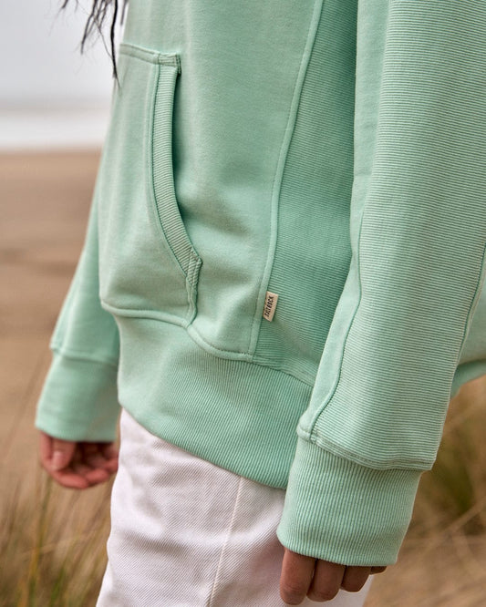 A woman wearing a Saltrock Shelley - Womens Pop Hoodie in Light Green on a beach.