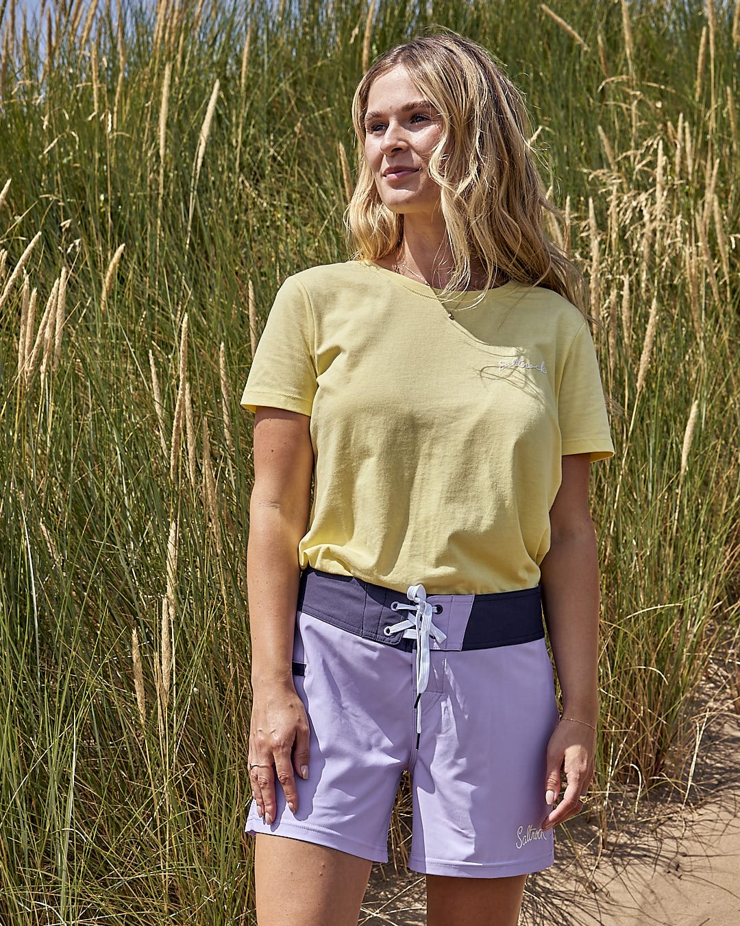 A woman in a Saltrock Shoreline - Womens Boardshort - Light Purple t-shirt standing in a field of tall grass.