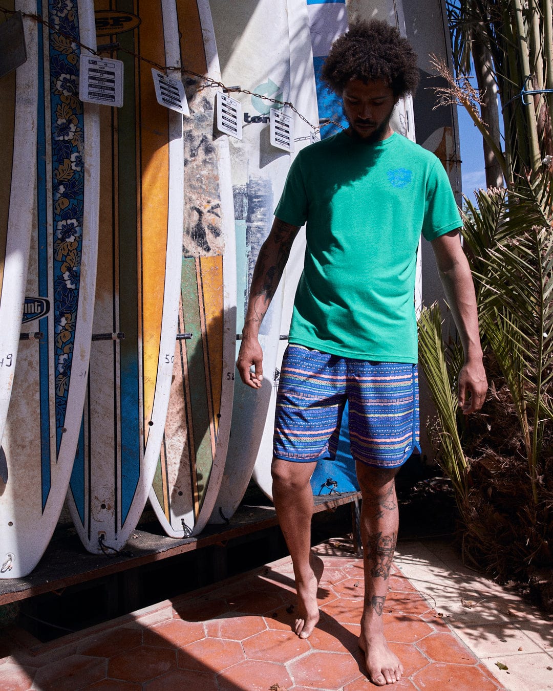 Man in green t-shirt and blue Silas boardshorts made of Repreve recycled material, standing next to a rack of surfboards under bright sunlight.