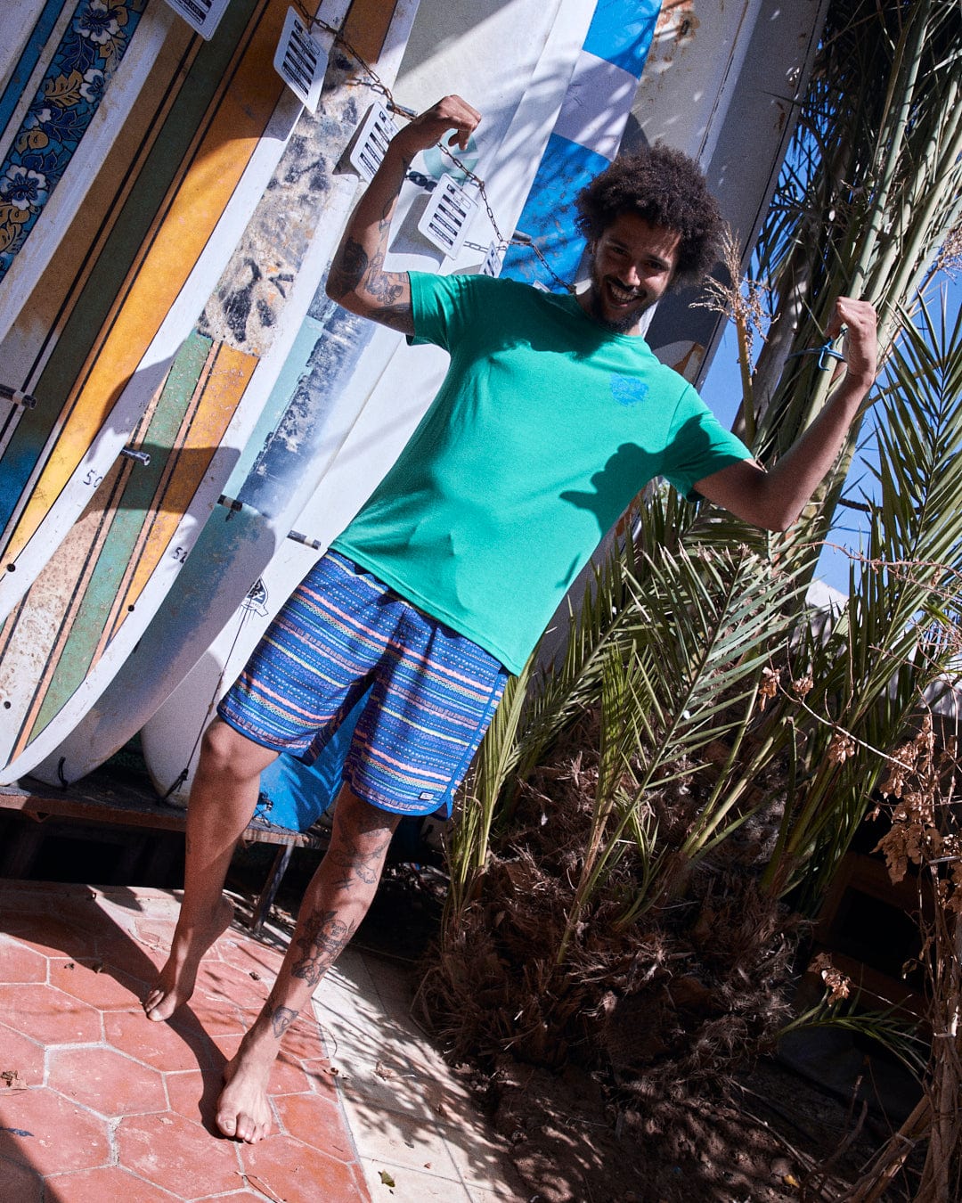 A joyful man in a green shirt and Saltrock Silas Mens Boardshorts in Blue Stripe made from Repreve recycled material jumps excitedly near a rack of surfboards and palm plants.