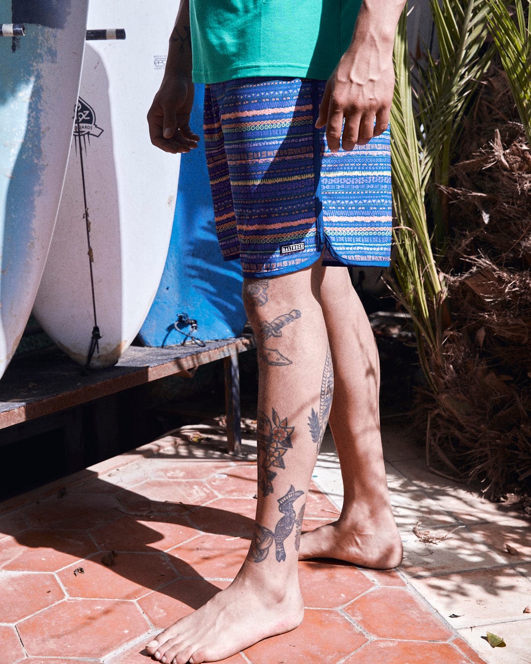 A person standing next to Saltrock surfboards, wearing blue Aztec print Silas boardshorts and displaying visible leg tattoos.