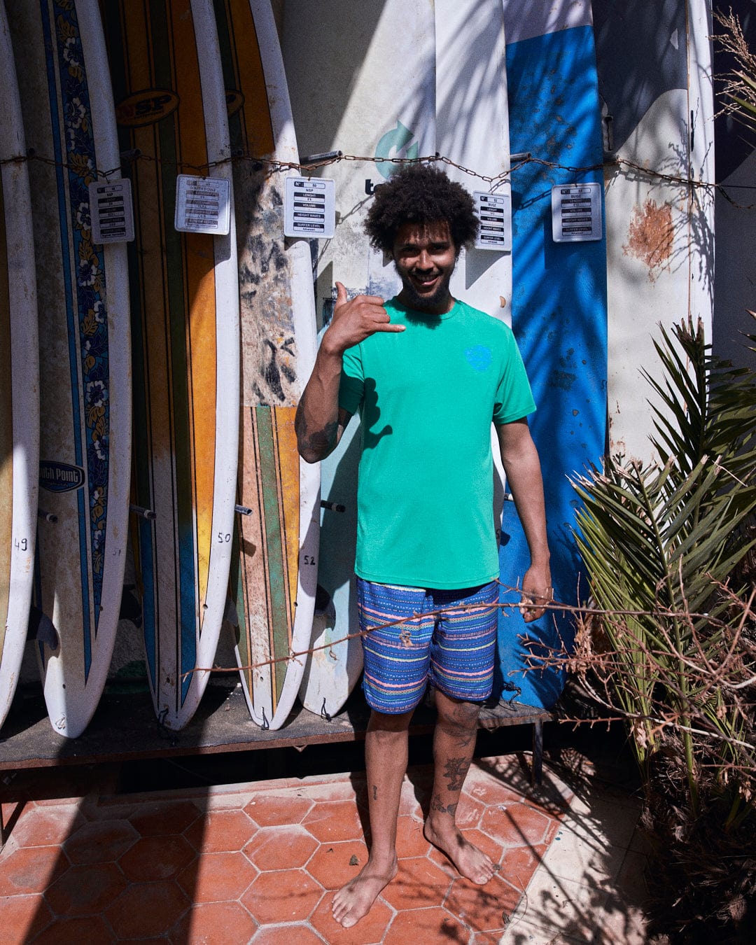 Young man with curly hair standing barefoot by a rack of colorful surfboards with Aztec print, making a shaka hand gesture, smiling in his Saltrock Silas Mens Boardshorts in Blue Stripe.