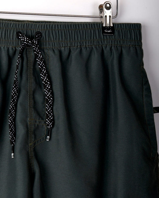 Close-up of dark green **Sinns - Mens Swimshorts - Green** from **Saltrock** with a peached soft hand feel, featuring black and white cord. They hang from a metal clip on a rack, showcasing their elasticated waist for added comfort.