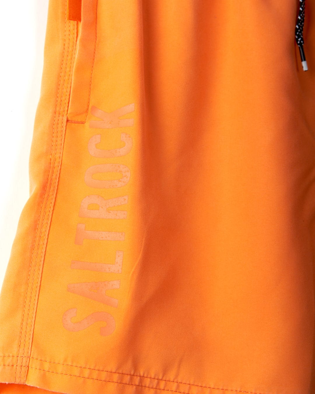 A close-up view of an orange fabric with "Saltrock" written vertically along the edge in a slightly faded white font. The edge features visible stitching, highlighting the distinctive Saltrock branding and peached soft hand feel of the Sinns - Mens Swimshorts - Orange.