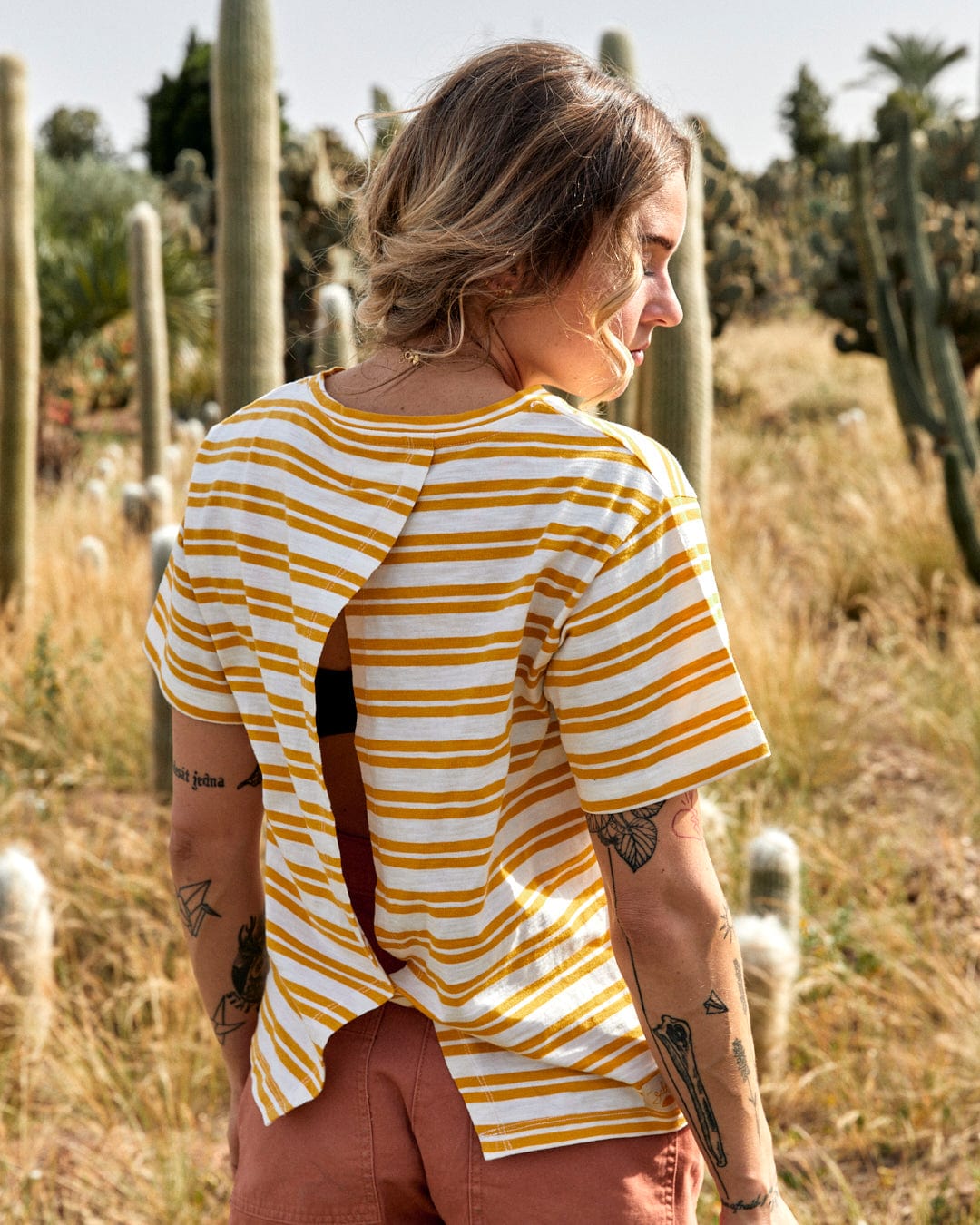 A woman in a Sky - Womens Short Sleeve T-Shirt - Yellow and white shirt and brown pants stands in a cactus field, seen from behind, looking to the side.