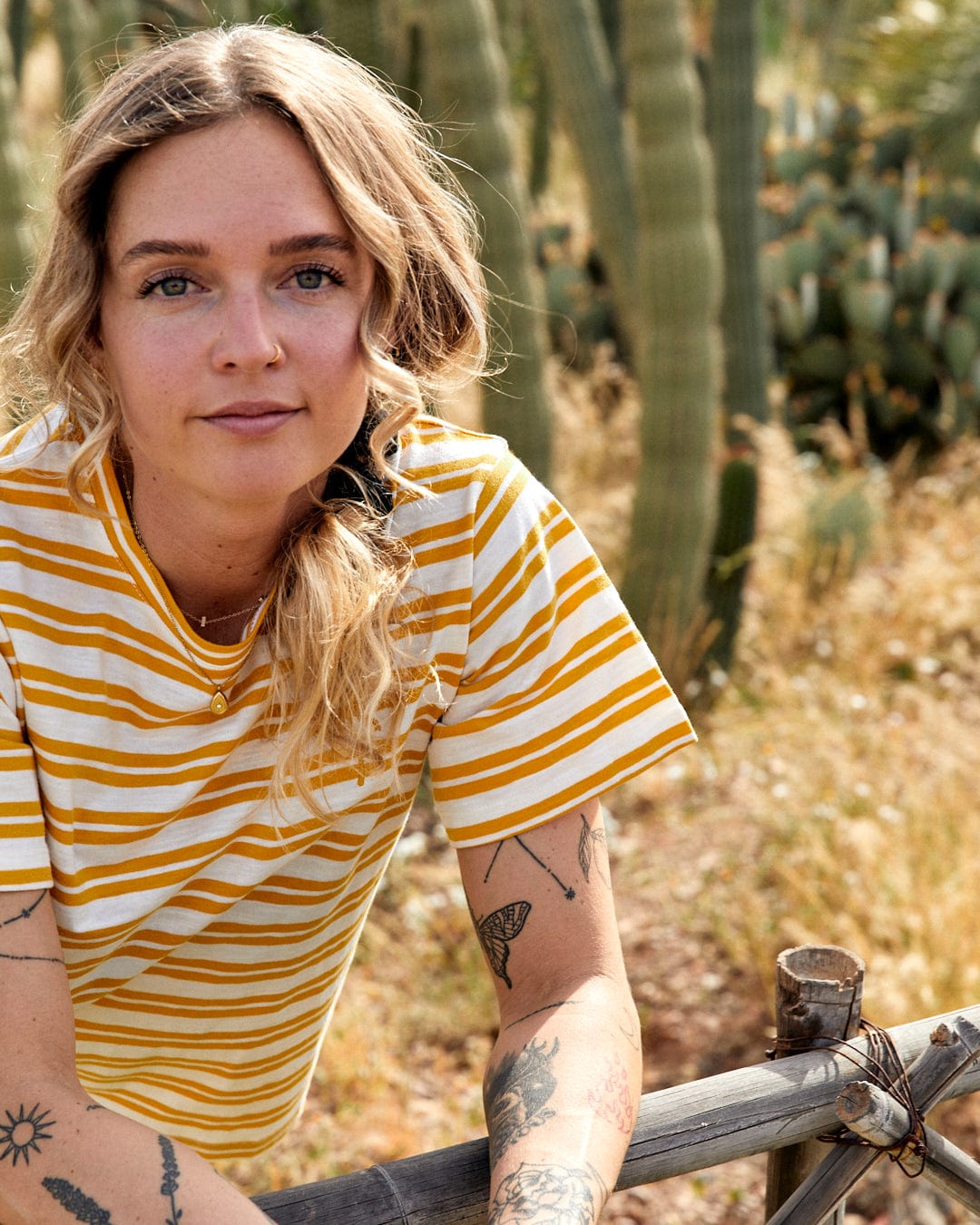 A woman with blonde hair, wearing a yellow Saltrock Sky - Womens Short Sleeve T-Shirt with yarn dye stripe print, leans on a wooden fence in front of tall cacti, looking at the camera.