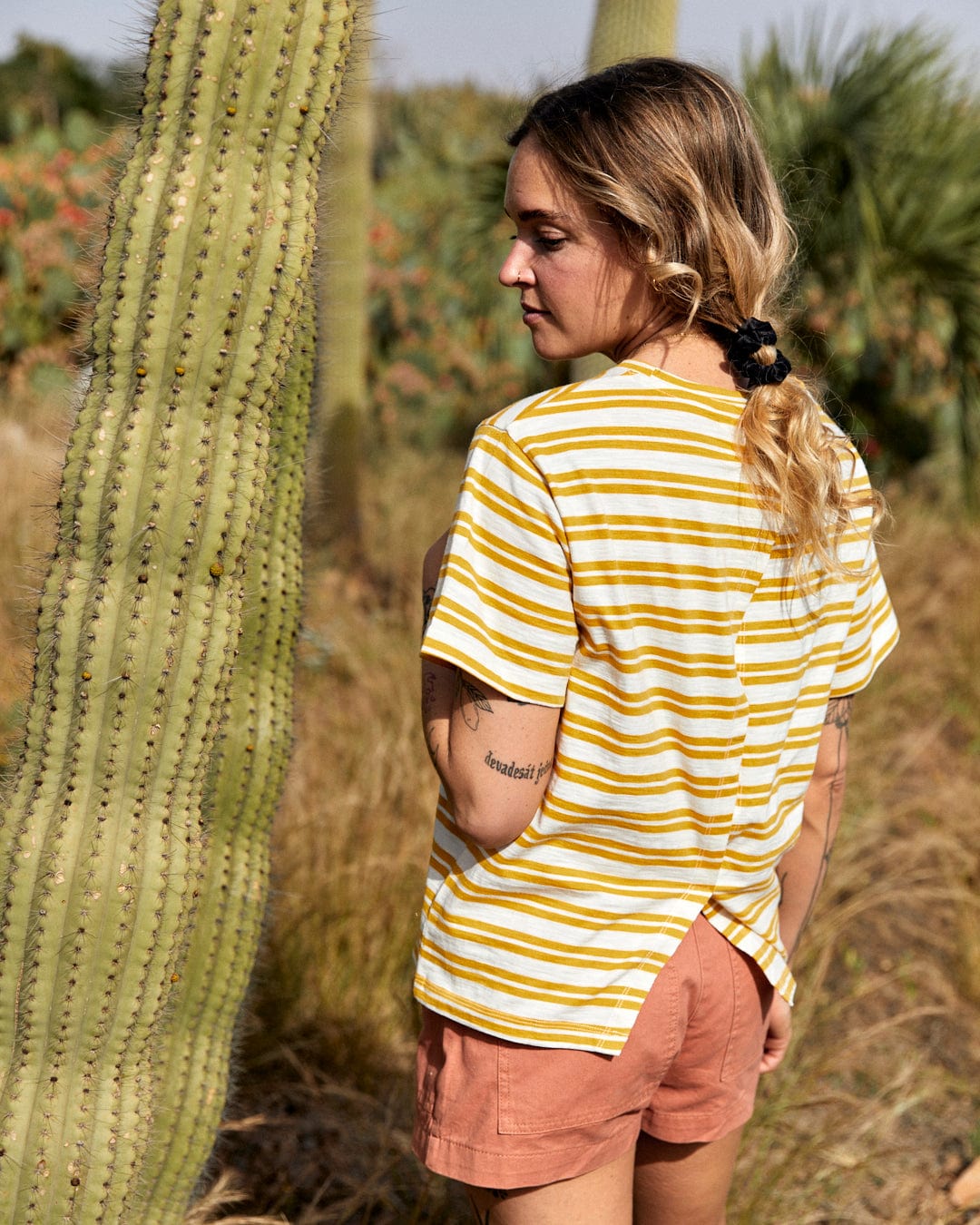 Woman in a Saltrock Sky - Women's Short Sleeve T-Shirt - Yellow standing next to a tall cactus in a sunny outdoor setting.